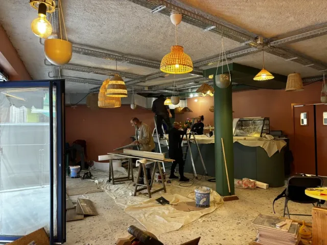 Fin de chantier dans la salle : réglage des luminaires sur escabeau, découpe sur tréteaux, murs terracotta et poteau vert déjà peints.