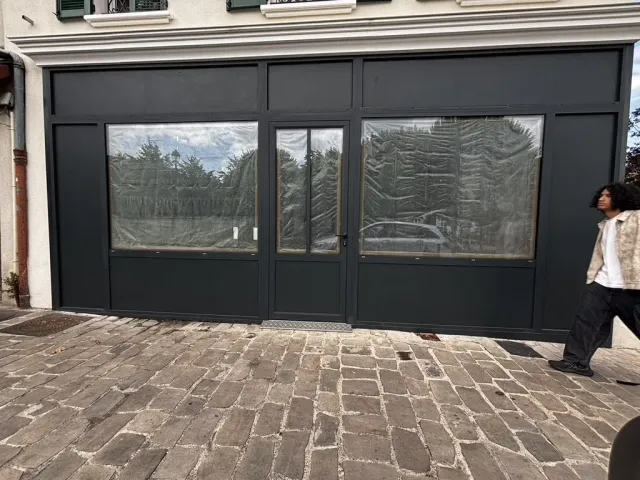 Devanture du local avant travaux, menuiserie anthracite neuve et vitrines occultées par des bâches, sur une rue pavée.
