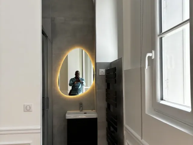 Salle d’eau créée dans une alcôve : miroir rétroéclairé, meuble suspendu noir, sèche-serviettes et sol en carreaux à motif géométrique.