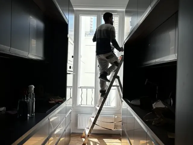 Cuisine en couloir aux façades sombres, réglage d’une menuiserie en contre-jour devant la fenêtre.