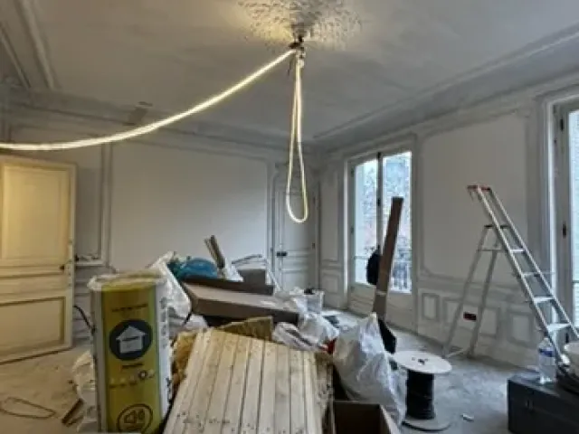 Séjour en chantier sous rosace de plâtre conservée, lames de parquet en attente de pose et touret de câble au sol.