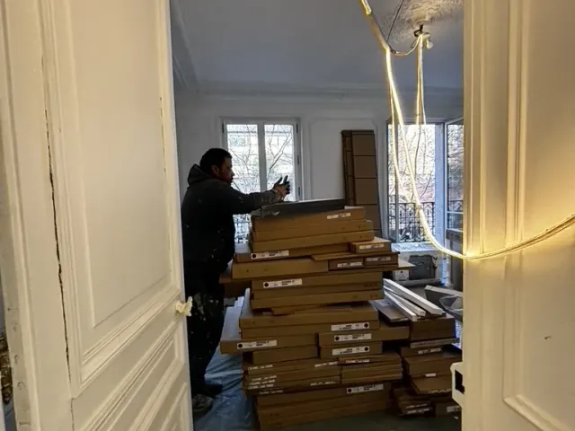 Livraison de parquet stockée en pile dans une pièce en travaux, éclairage provisoire en guirlande au plafond.