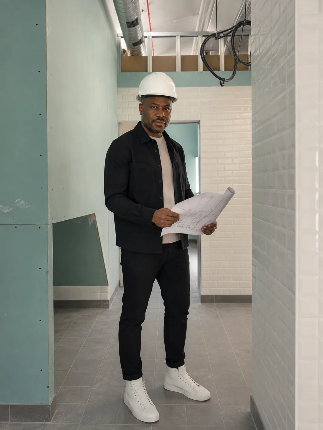 Charles Eschylle sur un chantier en cours, casque blanc et plans à la main, entre des cloisons en plaques de plâtre et une faïence métro blanche.